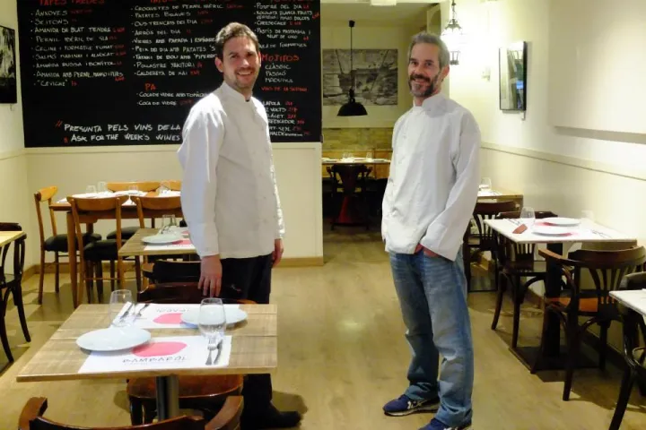 Two chefs in white jackets in the dining room, among the set tables and a large chalkboard.