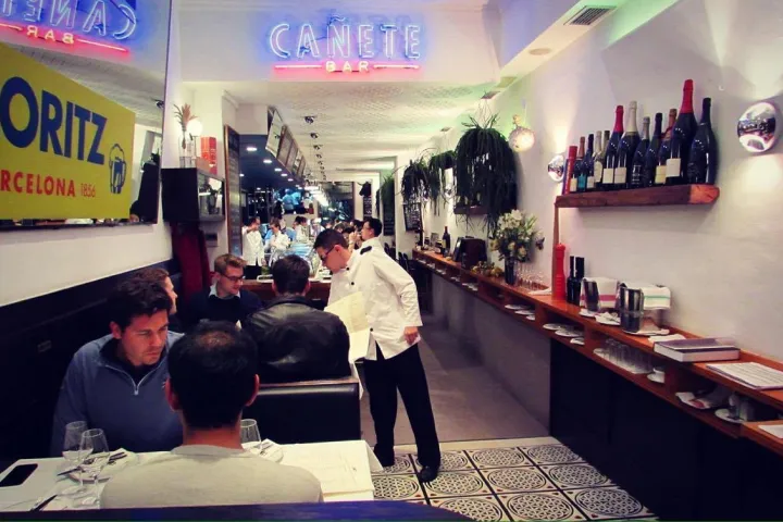 Patrons seated in a narrow room, with service and a row of bottles on the wall.