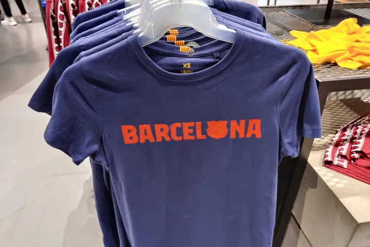 Ein marineblaues T-Shirt auf einem Bügel, mit dem Wort Barcelona und einem roten Wappen auf der Brust gedruckt.