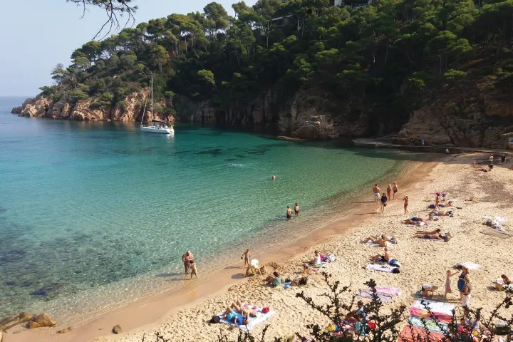 Der Sandstrand von Aiguablava erstreckt sich entlang türkisfarbenem Wasser, unterhalb einer bewaldeten Klippe, mit einem Segelboot vor Anker.