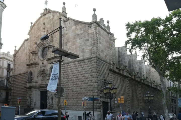 Il lungo lato in pietra della Chiesa di Betlem che costeggia la Rambla, tra i platani e i passanti