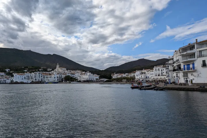 The bay of Cadaqués hugs the white houses along the waterfront; the Santa Maria church stands out above the rooftops under a cloudy sky.