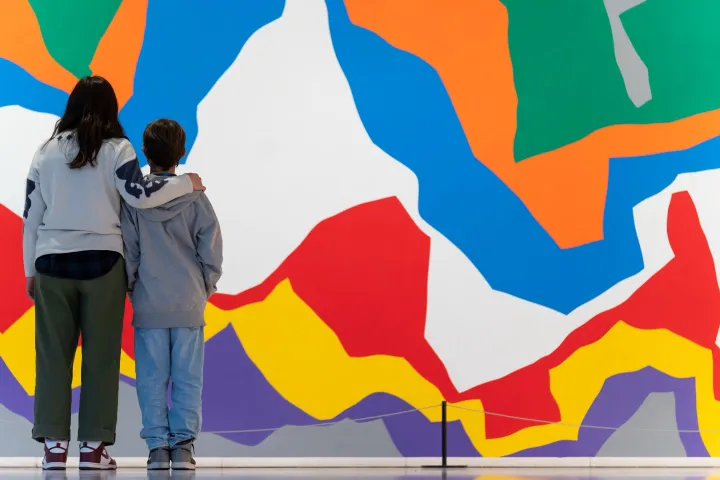 An adult and a child, seen from behind, with an arm around the shoulder, in front of a large mural with bold, vibrant colors