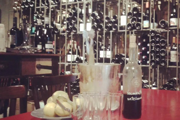 Gläser, Flaschen und ein Eiskübel auf einem roten Tisch vor Weinregalen.