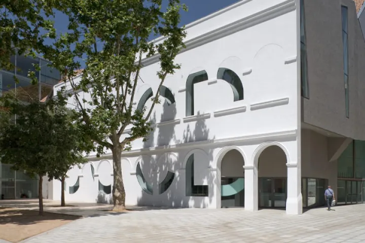 The white façade of the CCCB, featuring large arched openings, behind the plane trees of the square