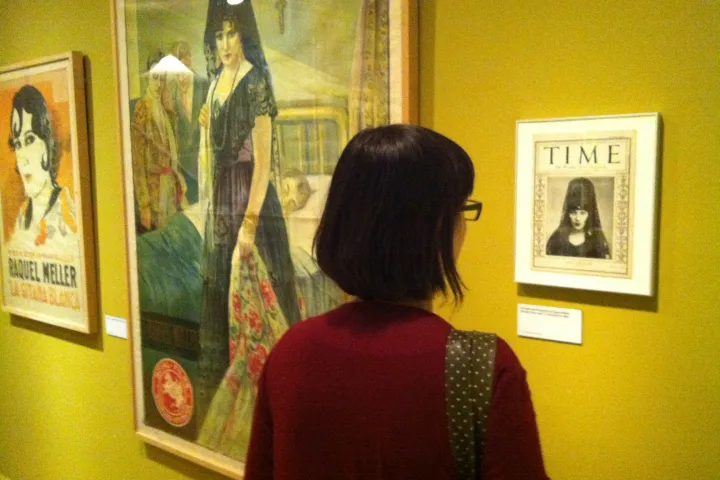 A visitor from behind in front of a painting, an old poster, and a framed magazine cover, in a room at the CCCB with yellow walls