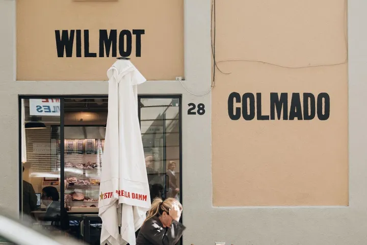 Facade of the colmado at number 28, featuring its display window, a parasol, and terrace tables.