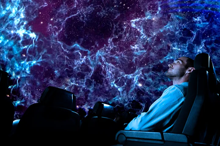Un visiteur allongé dans un fauteuil du planétarium, sous une projection de nébuleuse bleue et violette