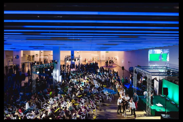 Uma grande sala com teto iluminado em azul, cheia de cadeiras e público, de frente para um palco e uma tela