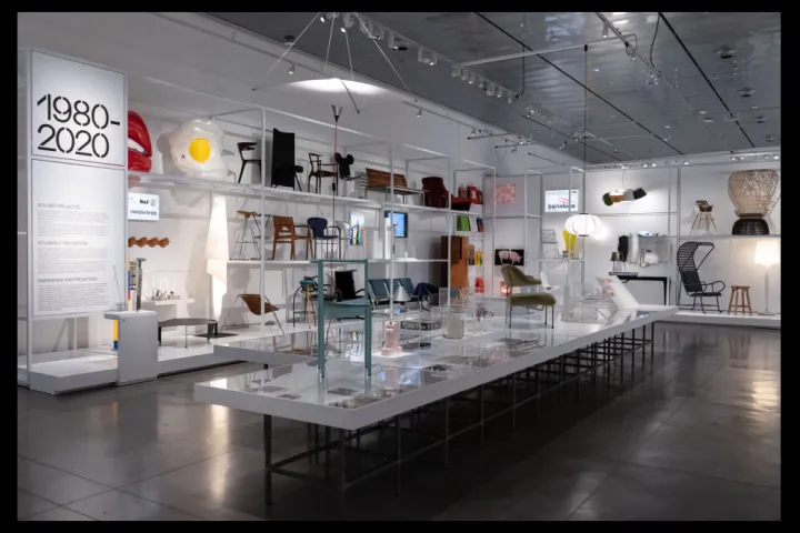 Cadeiras, lâmpadas e objetos de design apresentados em prateleiras brancas e uma longa vitrine baixa, sob o painel 1980-2020