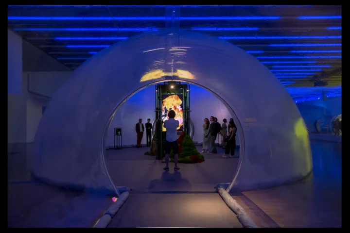 Uma estrutura inflável branca em forma de cúpula, iluminada de azul, na qual circulam visitantes