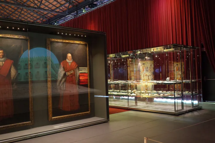 Ein Ausstellungsraum von El Born mit großen alten Porträts und einer Vitrine mit Geschirr vor einem roten Vorhang