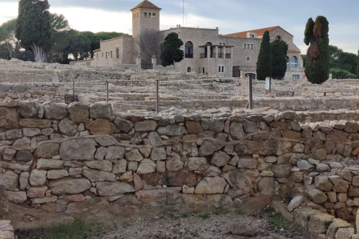 Die Steinmauern der archäologischen Stätte von Empúries stehen im Vordergrund vor dem Museumsgebäude und den Zypressen.