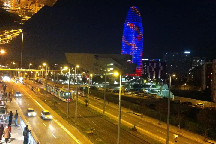 Torre Glòries illuminated as seen from the upper level of Encants Market, with the avenue and tram below