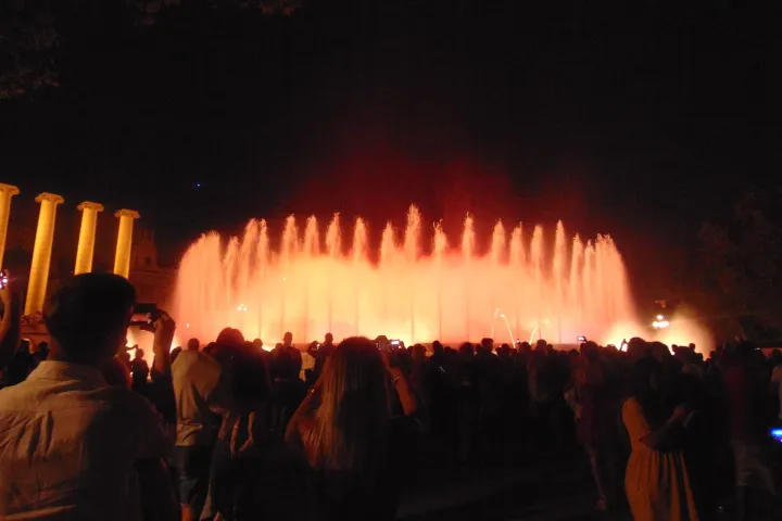 Spectators filming the orange jets of the fountain from behind, with the four illuminated columns on the left