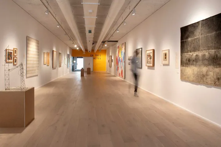 Eine lange Galerie mit hellem Parkett, gesäumt von aufgehängten Kunstwerken, durchquert von einer sich bewegenden Silhouette