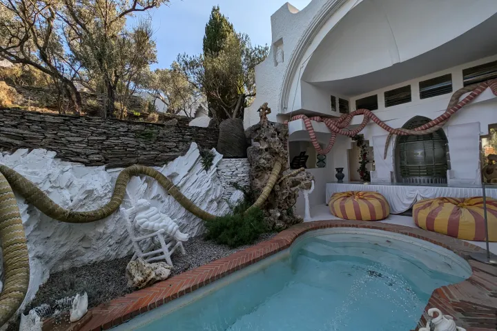 Un bassin aux bords arrondis longe un patio blanc, décoré de serpents sculptés et de coussins jaunes et rouges.