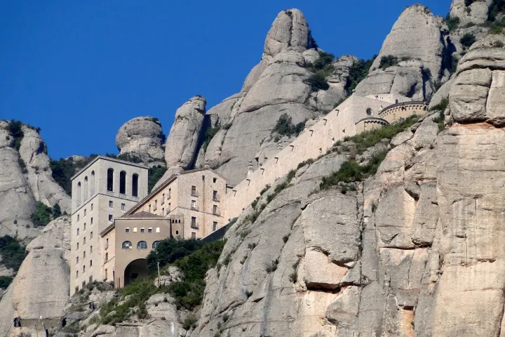 The buildings of Montserrat Abbey rise along the mountainside, beneath towering rock formations and a blue sky.