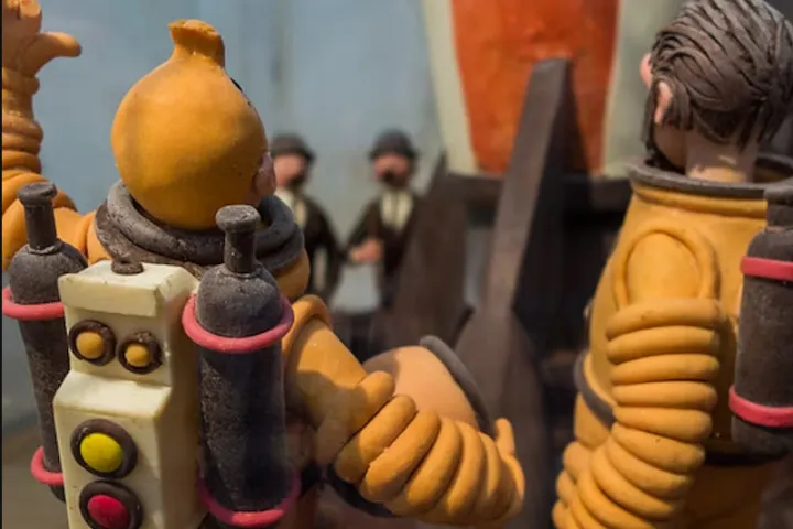 Personajes de cómic modelados en chocolate, con escafandra, frente a un cohete a cuadros rojos y blancos