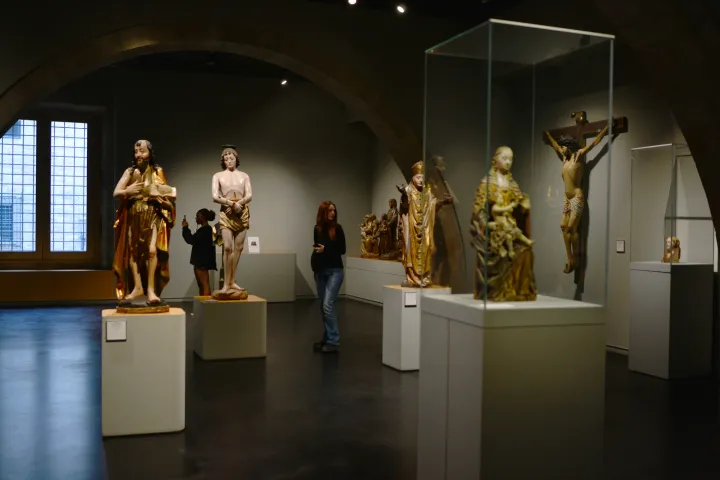 Esculturas religiosas policromadas em pedestais brancos, sob as abóbadas de pedra de uma sala do museu