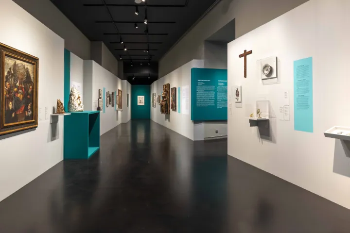 Uma galeria com paredes brancas e turquesa, onde convivem uma grande pintura religiosa, esculturas e ex-votos