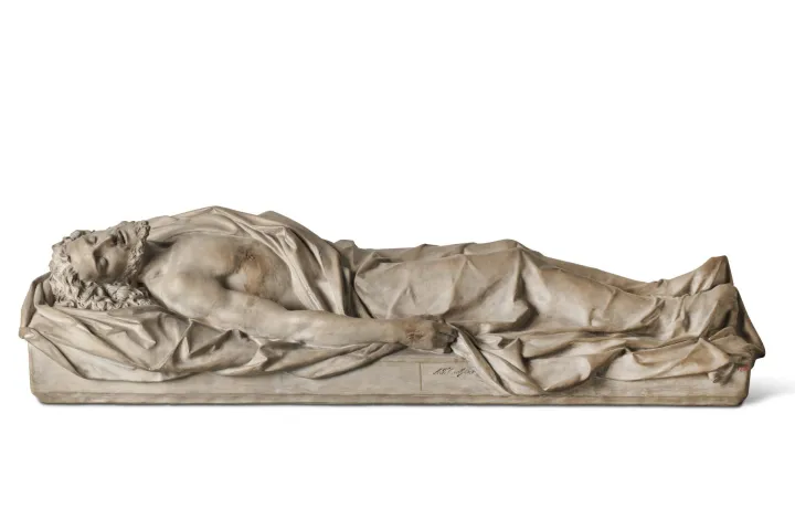 Uma escultura em pedra clara de um Cristo deitado, envolto em um sudário, sobre fundo branco