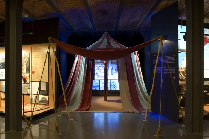 Une tente médiévale rayée rouge et blanc reconstituée sous les voûtes de brique du musée, au milieu des panneaux d'exposition
