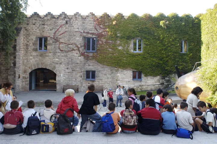 Un grupo de niños sentados frente a la fachada de piedra de las Drassanes, mirando a su acompañante
