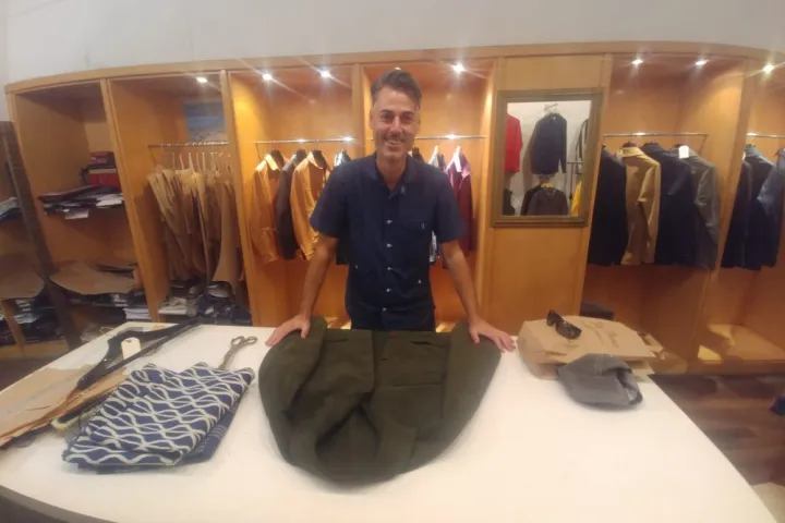 Un sastre detrás de una mesa de corte y un pantalón, frente a percheros de ropa.