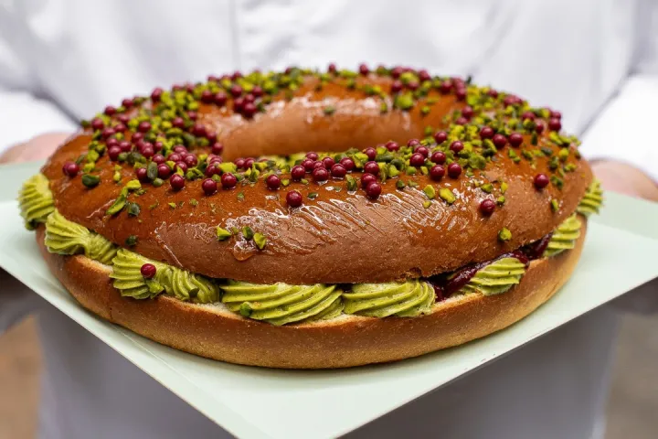 Una persona con atuendo de pastelero presenta un pastel en forma de corona adornado con crema verde.