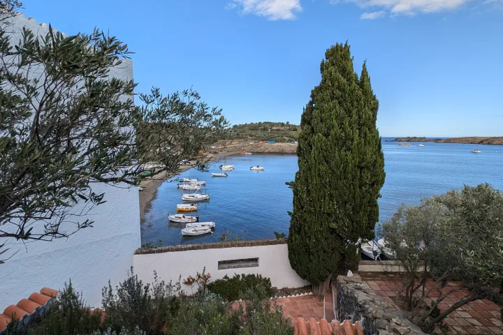Boats are moored in the bay of Portlligat, seen behind the white walls, a cypress, and the olive trees of the Dalí house garden.
