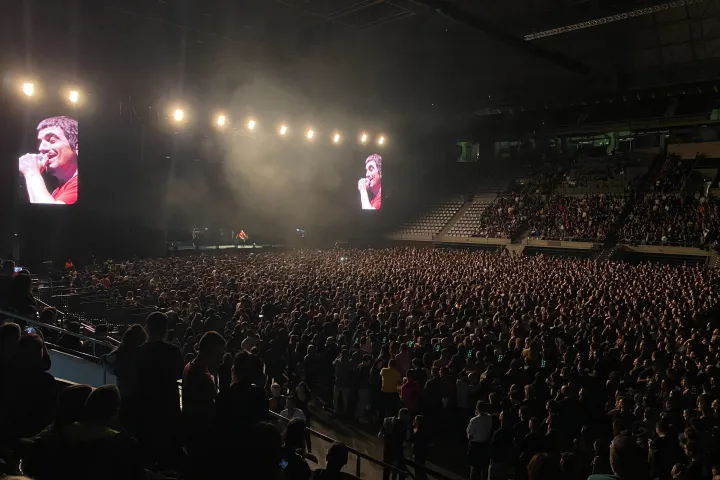 La sala immersa nel buio durante una serata di concerto, la folla in piedi e i maxi schermi ai lati del palco