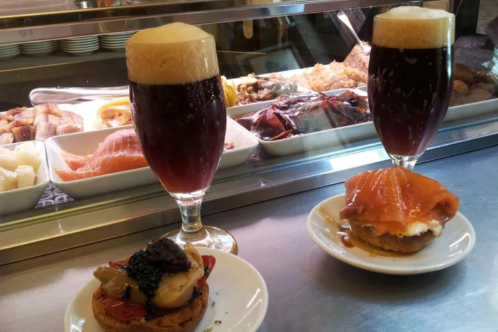 Two glasses of dark beer and montaditos on the counter, in front of the tapas display.