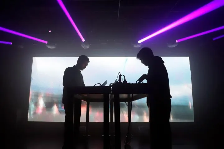 Dois músicos em suas máquinas no palco do Nitsa, sob feixes violetas.