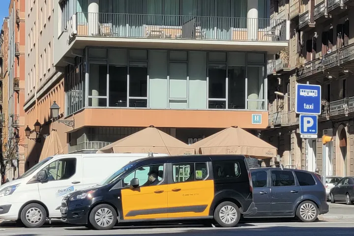 Taxi noir et jaune de type minivan devant un hôtel, près d’un panneau bleu indiquant une station de taxis.
