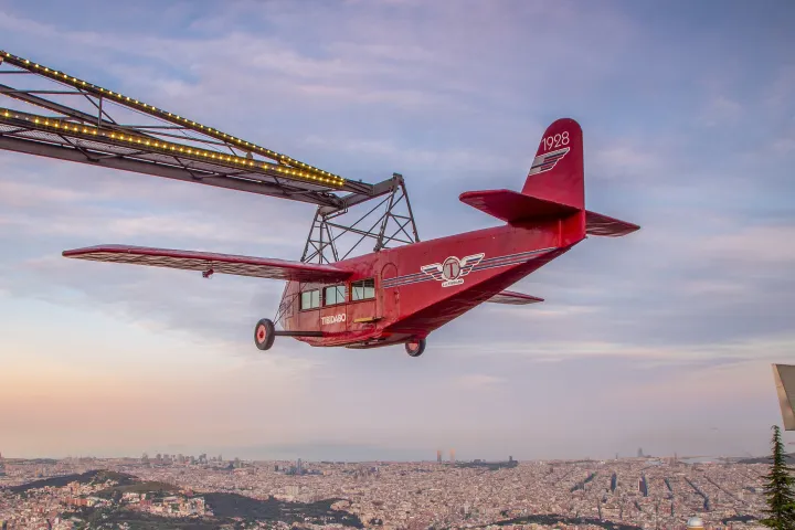 O avião vermelho suspenso em seu braço metálico, voando sobre Barcelona ao pôr do sol