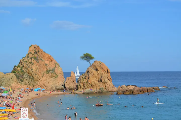 Die ockerfarbenen Felsen von Mar Menuda, von denen einer eine Kiefer trägt, überragen einen belebten Strand und die Badegäste.