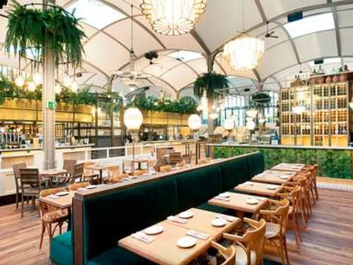 Salle d'El Nacional avec banquettes vertes, tables dressées, plantes suspendues et luminaires sous une voûte claire.
