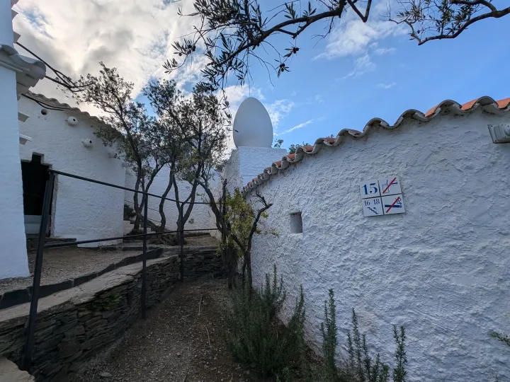 A gravel path runs along the white garden walls, under the olive trees, with a decorative egg above the roof.
