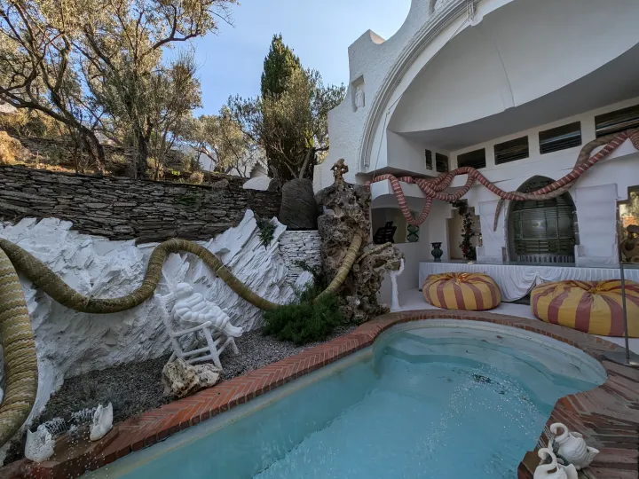 A rounded-edge pool runs alongside a white patio, adorned with sculpted snakes and yellow and red cushions.