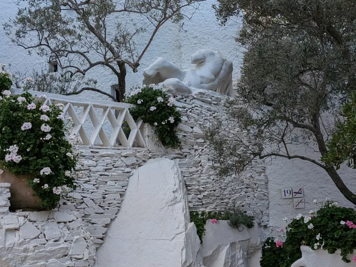 A white stone staircase ascends between olive trees, flower pots, and a crisscross railing.