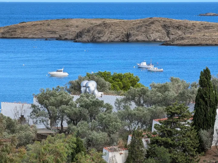 The white walls and olive trees of the garden overlook the bay of Portlligat, where several boats are anchored.