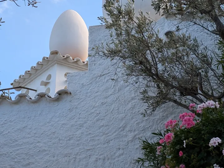 A monumental white egg rises from the roof, above a white wall lined with pink flowers and olive branches.