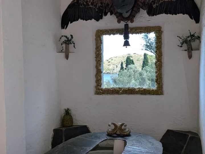 A table and curved benches occupy a white room, beneath a mirror and a taxidermied bird with outstretched wings.