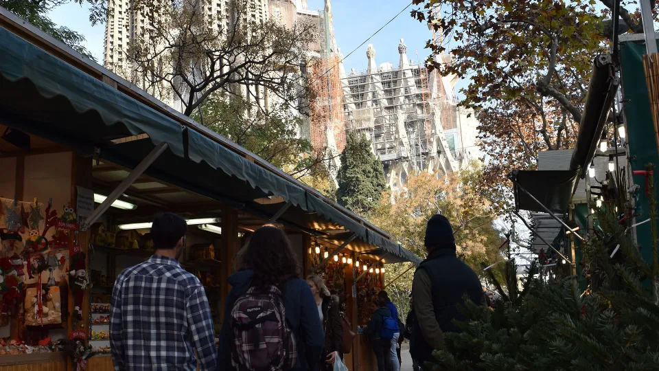 Ein Paar spaziert zwischen den Ständen der Fira de Nadal, die Türme der im Bau befindlichen Sagrada Família ragen darüber auf