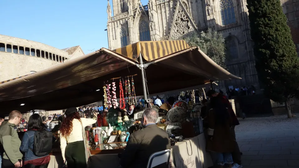 La fila di bancarelle del Mercat Gòtic e la folla di curiosi sulla Plaça Nova, davanti alla muraglia romana