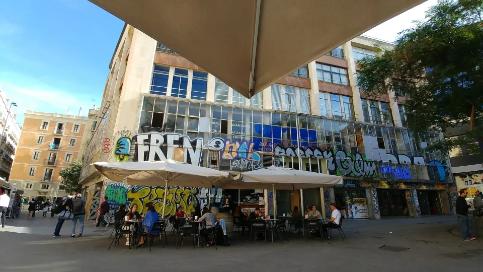 Uma varanda de café em uma praça do Raval, em frente a um prédio coberto de grafites