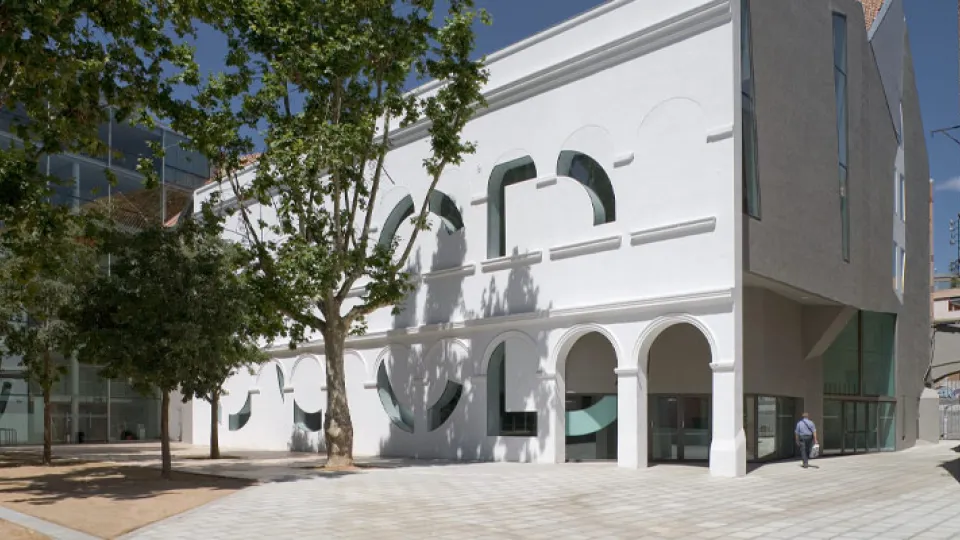 The white façade of the CCCB, featuring large arched openings, behind the plane trees of the square
