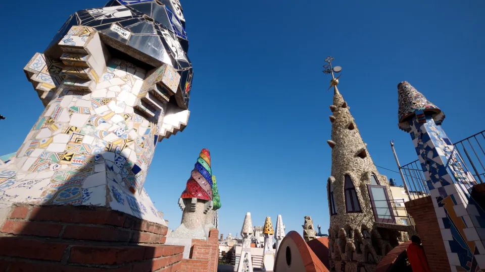 Die Dachterrasse des Palau Güell und seine mit buntem Trencadís verkleideten Schornsteine, um die zentrale Spitze, unter einem blauen Himmel