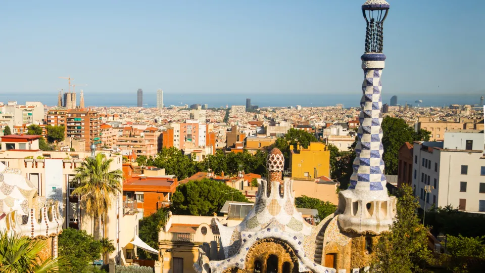 Les pavillons de l’entrée du Parc Güell et la flèche surmontée de sa croix, vus depuis la terrasse, avec Barcelone et la mer à l’horizon